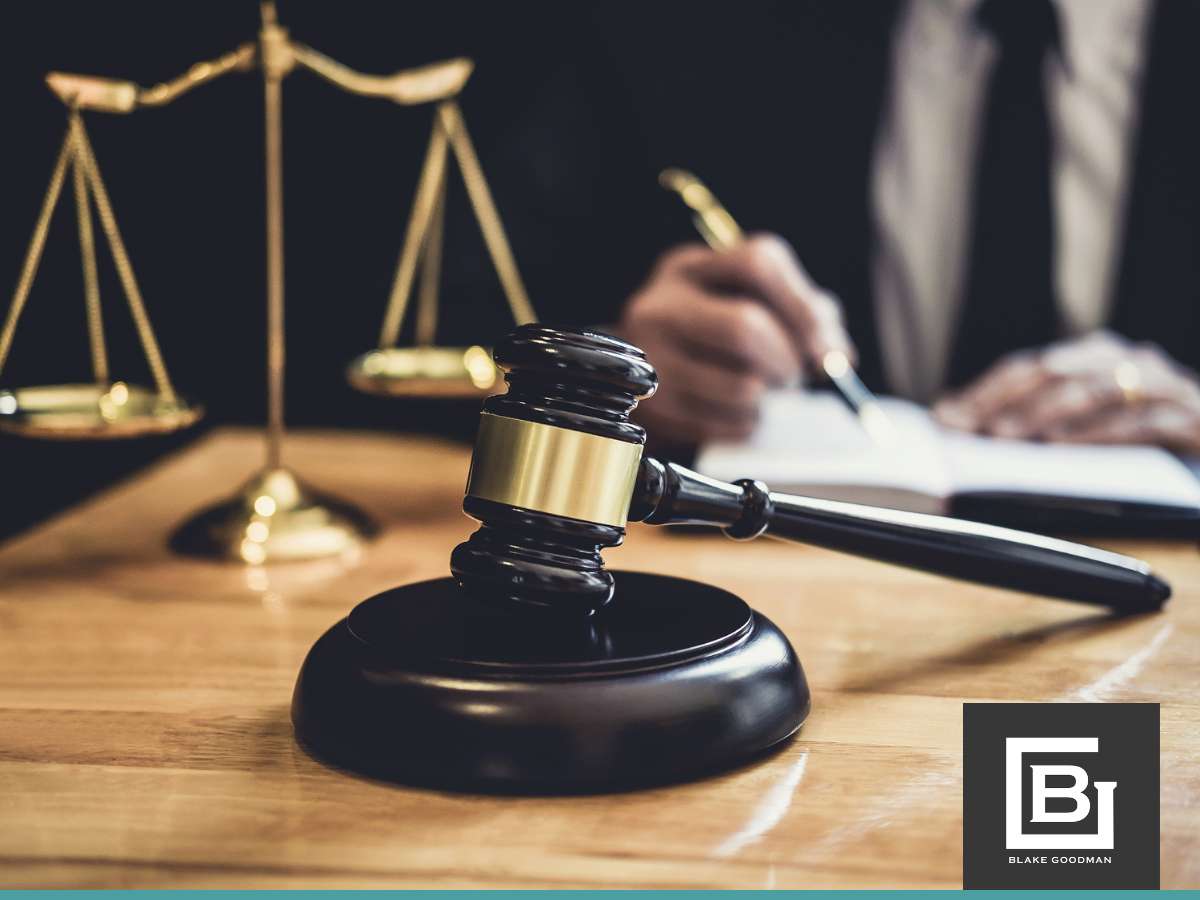 A professional setting with a wooden gavel on its base in the foreground, scales of justice in the background, and a person writing, possibly discussing loan modification vs. refinancing options.